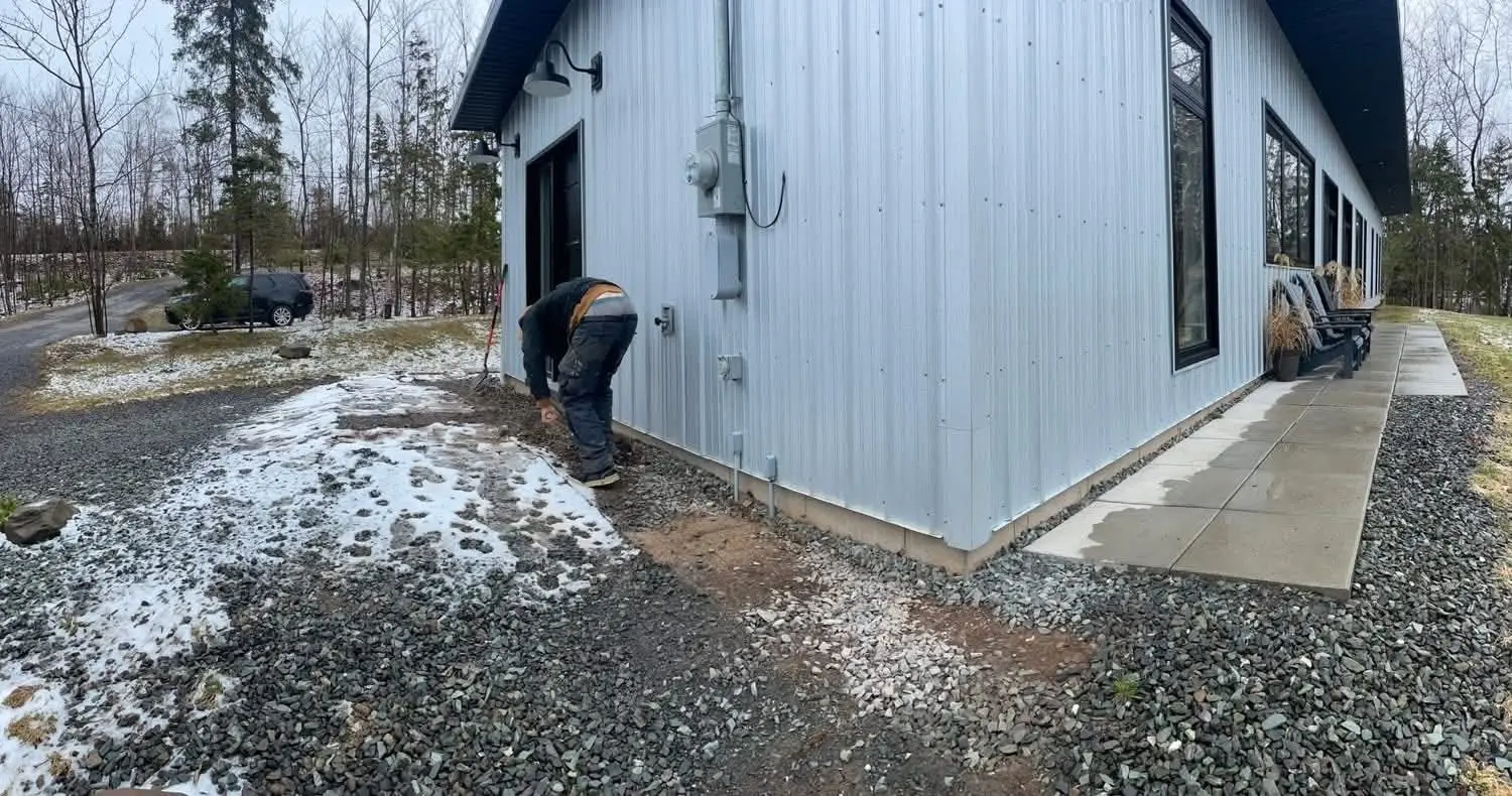 Crushed gravel base being prepared beside a metal-sided house in early winter